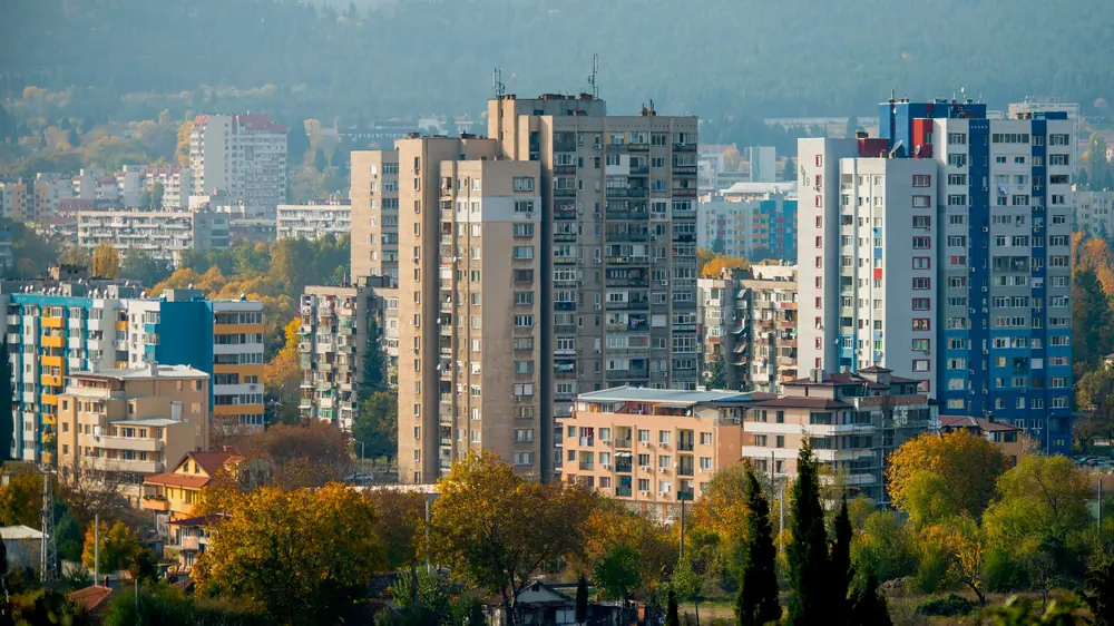 Високи жилищни блокове сред есенни дървета, зад тях — хълмове