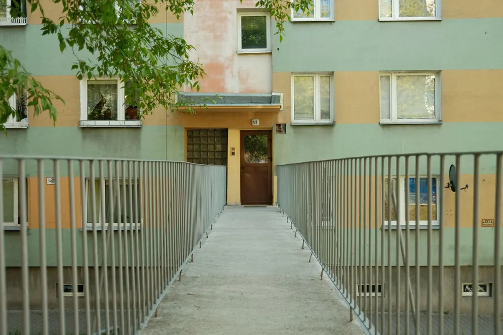 Пешеходна пътека с метални парапети към входа на жилищен блок с пастелна фасада и дървена врата